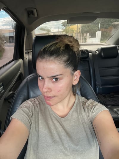 A woman with brown hair pulled back in a bun, wearing a grey shirt with silver embellishments on the sleeves, sits in the driver's seat of a car. The car has black leather seats and interior. Outside the window, the environment appears to be a street or residential area in Sumalao, Argentina, discernible by the architectural styles and vegetation. The image is taken from a slightly high angle, looking down at the woman. She is looking slightly upwards at the camera. A tattoo is visible on her left arm.