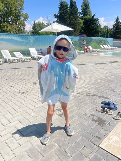 A young child wearing a shark-themed hooded towel and dark sunglasses stands on a paved walkway near a swimming pool in the city of Gagra, Abkhazia. The child is facing the camera, with their body angled slightly to the right. Several white lounge chairs are visible in the background, along with a decorative blue tarp depicting a landscape. In the distance, other people can be seen relaxing by the pool. The ground is made of gray interlocking paving stones, with some damp spots and shadows cast by the sun. A pair of dark blue slides and two camouflage-patterned slides are placed on the ground to the right of the child. The sky is bright blue with scattered white clouds, and lush green trees line the perimeter of the pool area.