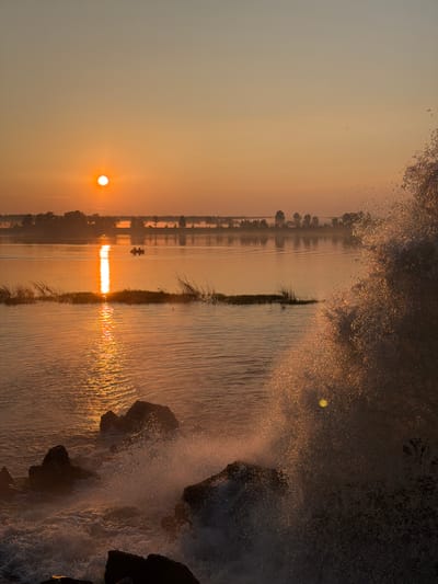 At sunrise, the sun, a bright orb, is positioned high in the pale orange sky. Its reflection creates a shimmering path across the calm water. In the distance, a treeline marks the shore, with a thin layer of mist visible between the water and the trees. Two people are in a small boat in the middle of the water, moving towards the shore. In the foreground, large, dark rocks are partially submerged in the water, with waves crashing against them, creating a spray of water droplets that catch the sunlight. The overall scene is one of tranquility and natural beauty, captured at the beginning of a new day. The location is the bank of the Kama River in the city of Chernushka, Russia.
