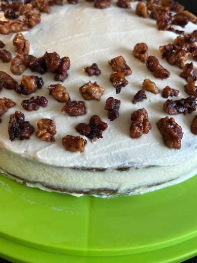 The image shows a close-up, overhead view of a frosted cake, likely a carrot cake due to its prominent topping of glazed walnuts. The cake is round and has been sliced, revealing layers of dark cake and white frosting. The frosting is applied unevenly, giving it a rustic appearance. Scattered across the top of the cake are numerous pieces of chopped walnuts, many of which appear to be candied or glazed, giving them a shiny, rich brown color. The cake is positioned on a bright, lime green plate or platter. The lighting suggests it is daytime, possibly indoors given the uniform illumination and lack of shadows. The setting appears to be a kitchen or dining area. There is no visible text in the image. The overall impression is of a homemade, indulgent dessert.