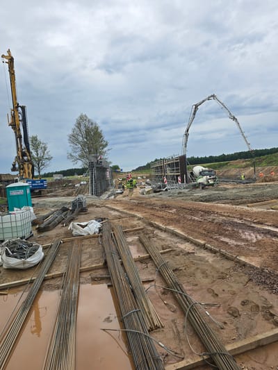 This image depicts a construction site in Piecnik, Poland. The scene is dominated by a large excavation with several ongoing construction activities. In the foreground, numerous bundles of rebar are stacked, some partially submerged in muddy puddles, indicating recent rain or poor drainage. A large yellow drilling rig is positioned on the left side of the frame.
Further into the site, a concrete pump with its long boom extends into the air, positioned over a foundation area where construction workers, wearing high-visibility vests and helmets, are actively working. A concrete mixer truck is visible near the pump, suggesting concrete pouring is in progress or has recently occurred. Scaffolding surrounds parts of the foundation structure, and a building material supplier's portable toilet, with the text "WESER.COM 601 715 009", is also present.
In the background, a dense line of trees borders the site, and the sky is filled with scattered clouds. The overall impression is one of a busy, large-scale construction project. The time of day appears to be daytime, possibly late morning or afternoon, given the lighting. The presence of mud suggests recent or ongoing inclement weather.