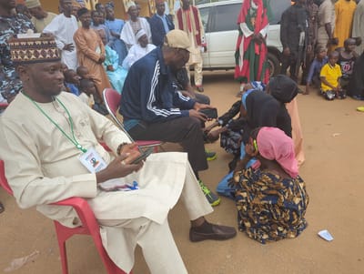 The image, taken in Kafanchan A, Nigeria, shows a gathering of people outdoors. A man wearing a traditional beige outfit and a patterned hat sits on a red plastic chair, holding a cell phone. Another man in a blue jacket and baseball cap sits nearby, also holding a cell phone. Several children are seated on the ground. Some are interacting with the man in the blue jacket. Adults dressed in both Western and traditional Nigerian clothing stand in the background. A white vehicle and armed security personnel are visible in the background. The ground is a reddish-brown color.