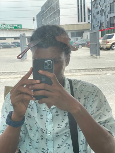 A person is taking a selfie with a black iPhone in front of a mirror, reflecting a street scene in Ojomu, Nigeria. The person is wearing a light blue shirt with a palm tree pattern and a blue watch on their left wrist. In the background, there is a green sign that reads "PRINCE EBENY SUPERMARKET" and several buildings and cars. The person is holding the phone with both hands, and their face is partially obscured by the phone and a comb that is positioned in their hair. The scene appears to be outdoors, possibly in a parking lot or an area adjacent to a street.