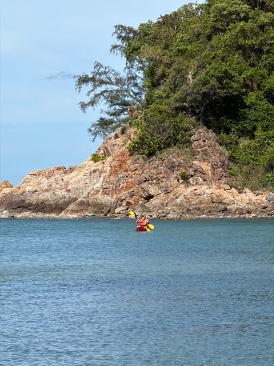 This is a vertical, medium-shot photograph taken outdoors. The scene features a rocky cliff face covered in lush green vegetation on the left, bordering a calm, light blue body of water on the right. The sky, a pale blue, occupies the upper left portion of the image, with some wispy white clouds visible.
In the water, near the base of the cliff, a red kayak is visible with two people inside. They are wearing orange life vests. One person is paddling with a yellow paddle, and another is seated behind them. The kayak is positioned close to the rocky shore.
The lighting suggests it is daytime, likely late morning or early afternoon, with clear weather. The overall mood is peaceful and serene, highlighting the natural beauty of the coastal landscape. The location is identified as Baan Plai Laem, Thailand. There is no visible text in the image.