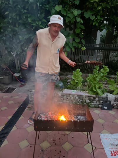 A man wearing a white bucket hat and a light-colored sleeveless shirt with camouflage shorts stands over a metal barbecue grill in a yard. He is holding a grill basket with food over the flames. Smoke rises from the grill and the surrounding area. The ground is paved with reddish-pink and yellow tiles. In the background, there is dense green foliage, a wooden fence, and some potted plants. The overall impression is a casual outdoor cooking scene. The location is identified as the city of Gagra, Abkhazia.