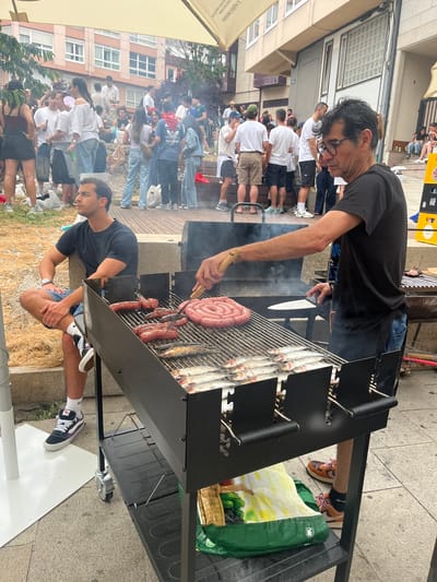 In A Coruña, Spain, an outdoor scene unfolds with a barbecue as the focal point. A man with glasses, wearing a black t-shirt and jeans, is actively grilling. He is using a spatula and a knife, and the barbecue is laden with several sausages, one coiled into a spiral, and numerous small fish. Smoke rises from the grill, indicating the cooking process.
In the foreground, another man is seated, wearing a dark t-shirt and denim shorts. He is looking to his left, seemingly observing the surroundings. He has a watch on his left wrist and is wearing sneakers.
Behind the barbecue, a crowd of people are gathered, creating a lively atmosphere. They are dressed casually, suggesting a social event or a public gathering. Some are standing, while others are seated on tiered seating structures. The backdrop consists of buildings with balconies and windows, typical of an urban setting. The ground is paved, and some areas have scattered dry grass or foliage. A yellow umbrella or canopy is partially visible overhead, providing shade. The overall impression is of a community event or a casual get-together in a city environment.