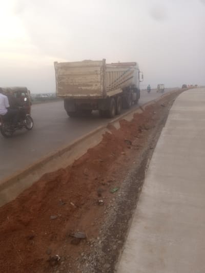 The image shows a dump truck driving on a road under construction in Goni Gara, Nigeria. The truck is carrying dirt, and there is a trench next to the road filled with dirt and gravel. Other vehicles, including a motorcycle and a tricycle, are also visible on the road. The sky is overcast, and the overall lighting suggests it is either early morning or late afternoon. The road appears to be newly paved in some sections.