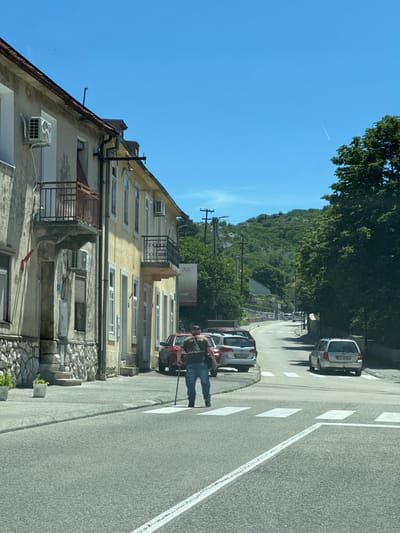 The image depicts a street scene in Cetinje, Montenegro, on a clear, sunny day. A man, appearing to be elderly and using a cane for support, is crossing a pedestrian crossing. He is wearing a t-shirt and blue jeans. Several cars are parked along the street and on the road, including a silver station wagon with the license plate CT AK924. On the left side of the street, there is a building with a stone foundation and a yellow-painted upper story. This building has several windows, balconies, and air conditioning units visible. Trees line the right side of the street, and a hilly, tree-covered landscape forms the background. The road is paved, and the sky is a vibrant blue with a faint contrail from an aircraft.
