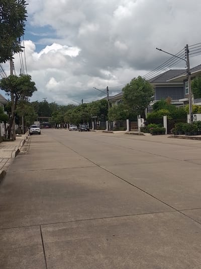 The image depicts a residential street in Si Sunthon, Thailand, under a cloudy sky. The street is paved with concrete and has visible expansion joints. On the left side of the street, a sidewalk with a concrete curb is present, bordered by a low white fence and some trees. On the right side, modern houses with dark tiled roofs and large windows are visible, with well-maintained gardens and potted plants. Several cars are parked along the street: a white SUV is parked on the left, a white sedan and a dark sedan are parked on the right. Power lines and streetlights are present along the street. The environment appears to be a quiet, suburban neighborhood.