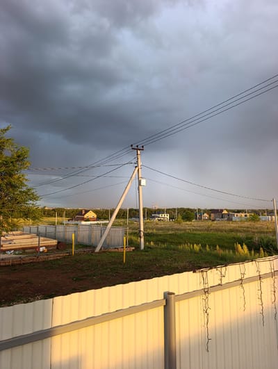 The image depicts a rural landscape in the city of Lenina, Russia, under a heavy, dark sky with storm clouds. In the foreground, a white corrugated metal fence is illuminated by golden sunset light, with icicle-like string lights hanging from its top edge. To the left, a construction site is visible, featuring stacks of lumber and construction materials next to a silver corrugated metal fence. Beyond this, a green field stretches out, dotted with yellow wildflowers. A prominent utility pole with multiple wires traverses the scene. In the middle ground, a series of houses with pitched roofs are scattered across the landscape, nestled amongst trees. In the background, a distant city skyline is faintly visible, partially obscured by haze and the dark clouds. The overall impression is one of an impending storm in a semi-developed rural area.