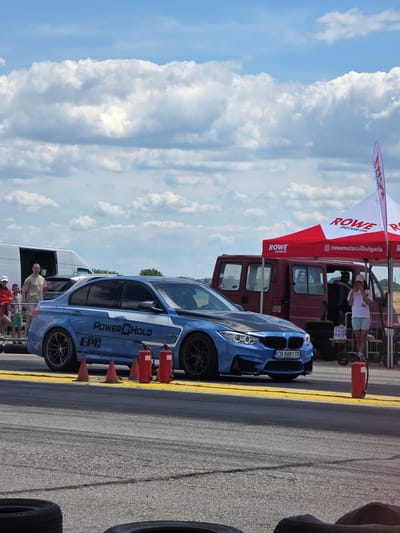 In Akatsievo, Bulgaria, on a sunny day, a blue BMW M4 Competition is parked on a asphalt track, a race or exhibition event. The car is adorned with the decals "Power HOLD" and "EPC". Beside the car, two red fire extinguishers stand upright on the yellow line of the track. Further back, a red van is parked near a red and white tent with the "ROWE MOTOR OIL" logo visible. Several individuals, including adults and children, are observing the scene. In the background, trees and a power line pole are present. The sky is filled with scattered white clouds.