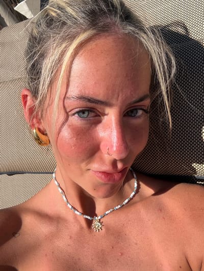 A woman lies on a dark, patterned surface. Her face is flushed from the sun, with light reflecting off her nose and forehead. She has light eyes, a nose ring, and wears a chunky gold earring. A beaded necklace with a sun pendant adorns her neck. Her blonde hair is streaked with grey. The picture is taken from a high angle, with the subject tilted slightly.