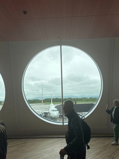 A man walks past a large circular window in what appears to be an airport terminal. The window overlooks a tarmac where a white airplane is parked, attached to a jet bridge. A red and white striped tower is visible in the background, along with trees and a cloudy sky. Another person, a woman, stands to the right of the window, holding a smartphone and taking a photo. The man in the foreground is wearing a dark jacket and backpack and is pulling a suitcase. The floor is made of light-colored wood planks. The ceiling is paneled with wood. The scene is located in Yuzhnyye Koryaki, Russia.
