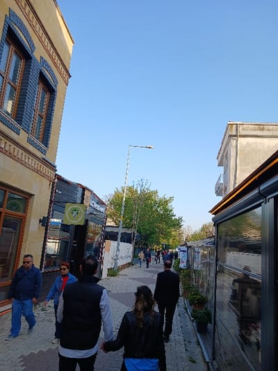 The image shows a narrow street in Istanbul, Türkiye, located at latitude 40.959 and longitude 28.823. The street is lined with buildings on both sides, with a yellow building on the left side. The street is paved with brick and is relatively empty. There are a few people walking down the street. One man is walking towards the camera on the left side of the street. A woman is walking in the opposite direction, closer to the camera. Three people are walking away from the camera, two men and a woman. The street is illuminated by a streetlamp and the sun. There is a sign for a restaurant or shop on the left side of the street.