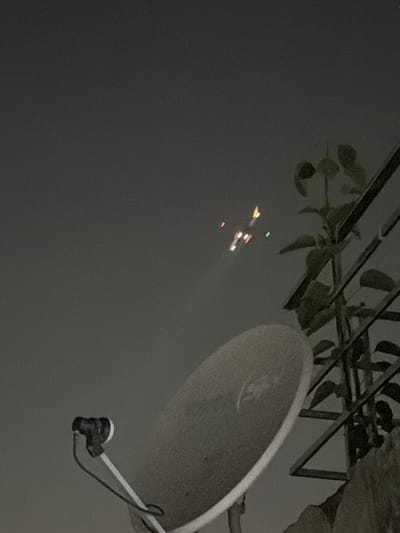 The media file shows an outdoor scene at night, likely in Delhi, India, given the context. The primary subjects are a satellite dish and a distant object with multiple lights in the sky. The satellite dish, in the foreground and to the right, is large and grey, with its LNB (low-noise block downconverter) and connecting wire clearly visible. Above and behind the dish, there are some dark, leafy branches, possibly from a tree or a plant, partially obscuring a metal structure that appears to be part of a railing or a canopy.

The most notable detail in the image is the collection of lights in the sky, appearing as a small, distant cluster. These lights are in various colors, including red, yellow, and green, and seem to be in motion or at different positions relative to each other. This cluster of lights could represent an aircraft, possibly a helicopter with its navigation lights illuminated, or perhaps distant fireworks, though the sharp, defined nature of the lights suggests an artificial source like an aircraft.

The overall ambiance of the image is dark and somewhat hazy, suggesting it might be nighttime or dusk, and potentially a humid or dusty atmosphere, common in Delhi. The image is captured from a low angle, looking upwards, which emphasizes the satellite dish and the sky. There is no visible text in the media file. The scene is static, with no clear indication of human activity directly present, beyond the implied presence of someone operating or maintaining the satellite dish.