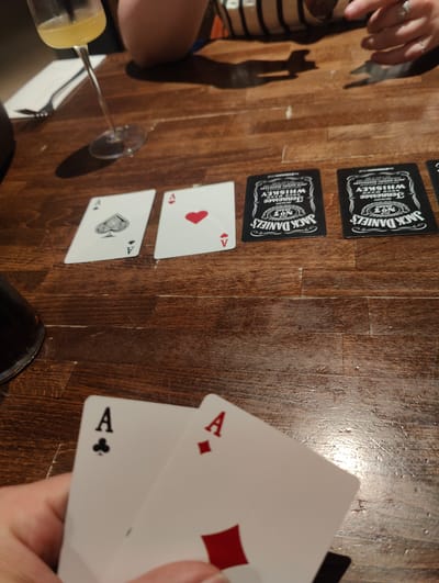 The image shows a close-up of a playing card game in progress on a wooden table. Five cards are visible, three face-up on the table and two held in a person's hand in the foreground. The table cards are: the Ace of Spades, the Ace of Hearts, and what appear to be two Jack Daniel's promotional cards (or potentially cards from a novelty deck). The person is holding the Ace of Clubs and the Ace of Diamonds. A glass of a yellow drink, a person wearing a striped shirt, silverware and a dark container are also visible in the background. The image does not provide any information to determine if the game is located in the city of East Suffolk, United Kingdom.