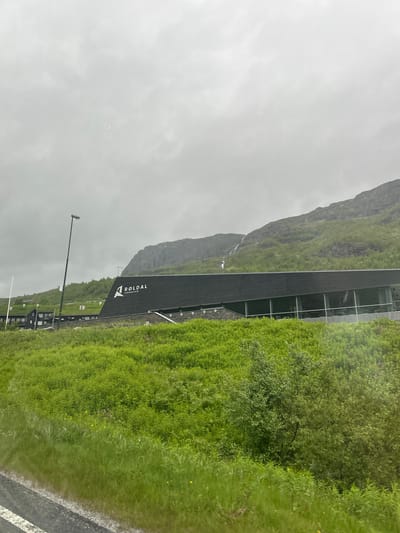 This media file displays a building with large glass windows situated in a lush, green, mountainous landscape. The building is dark-colored, possibly black or dark gray, and features the text "RØLDAL TERAPARKON" in white lettering with a triangular logo. Surrounding the building is vibrant green grass and foliage, indicating a thriving natural environment. In the background, a steep, rocky mountain rises, partially obscured by mist or clouds. A thin waterfall can be seen cascading down the mountainside. The sky is overcast, suggesting a cloudy or rainy day. A street lamp is visible in the foreground. The scene appears to be a modern architectural structure integrated into a dramatic natural setting, possibly a resort or a wellness center, given the "TERAPARKON" text. The overall mood is serene and slightly dramatic due to the imposing mountains and the overcast weather.
