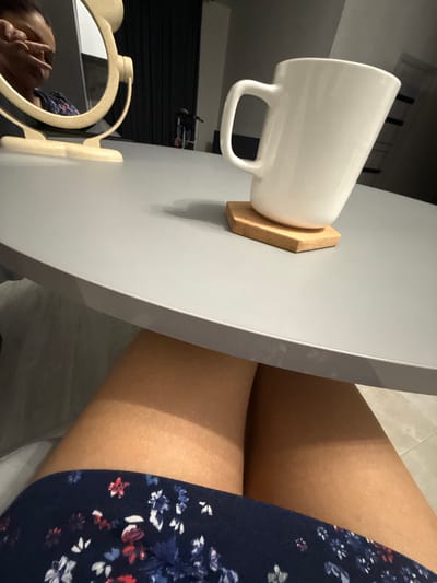 In this overhead shot, a white mug rests on a hexagonal wooden coaster atop a light gray table. The mug's handle is visible, and it is positioned to the left of the frame. To the right, a person's bare legs are visible, clad in a navy blue dress adorned with a floral pattern featuring red, white, and blue flowers. The lighting suggests it might be daytime, possibly indoors or in a shaded outdoor area, as there are distinct shadows cast on the person's legs. In the lower left corner, a vanity mirror with a cream-colored frame reflects a person's face, with one hand raised to their cheek. The background appears to be a room with dark vertical elements, possibly shelving or blinds, and a tiled floor. The overall mood is casual and relaxed.