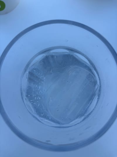 The image is a top-down close-up of a glass containing ice cubes and water. The glass is clear and has a thick rim. The ice cubes are large and appear to be melting, with water droplets visible on the inside of the glass. The lighting is dim and blueish, suggesting an indoor setting. There are no people or other objects visible in the image besides the glass and its contents.
There is no information within the image to suggest a location in Dénia, Spain.