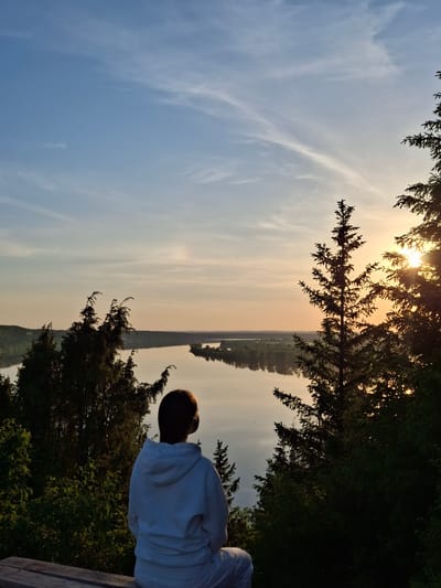 A person with short brown hair and a white sweatshirt sits facing a body of water from a wooden bench, framed by trees on either side. The sun is setting, casting a warm glow over the horizon in Прикамский, Russia. The sky is a gradient of blue to orange. A thin strip of land borders the water's edge, dotted with trees. The vegetation around the bench is lush and green.