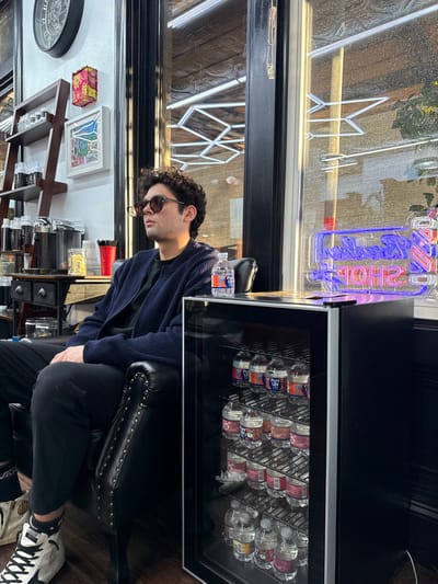 This image depicts the interior of a barbershop located in Boston, United States. The exact street address or specific neighborhood within Boston cannot be determined from the visual information provided.
A male individual with dark, curly hair, wearing dark sunglasses, a dark blue ribbed cardigan over a dark shirt, and black trousers, is seated in a black leather armchair with brass studding, facing towards a large window to his right. A bottle of Pure Life water rests on the armchair's right armrest. His left foot, clad in a black sock and a white and black high-top sneaker with a star emblem, is visible on the dark wood floor.
To the immediate right of the armchair, a compact black mini-fridge with a clear glass door is stocked with multiple rows of Pure Life bottled water. On top of the mini-fridge, a lit neon sign displays "BARBER SHOP" in blue and red.
In the background to the left, a dark wooden shelving unit holds various containers, likely haircare products. Below it, a dark wooden cabinet features a chrome dispenser, red plastic cups, and a clear container with yellow, possibly straw-like, items. The wall decor includes a large dark circular clock, a decorative red and gold rectangular box, and a framed colorful landscape artwork. Bright natural light enters the space through the large window, reflecting interior elements such as a patterned ceiling and another neon sign. The exterior view through the window is indistinct.