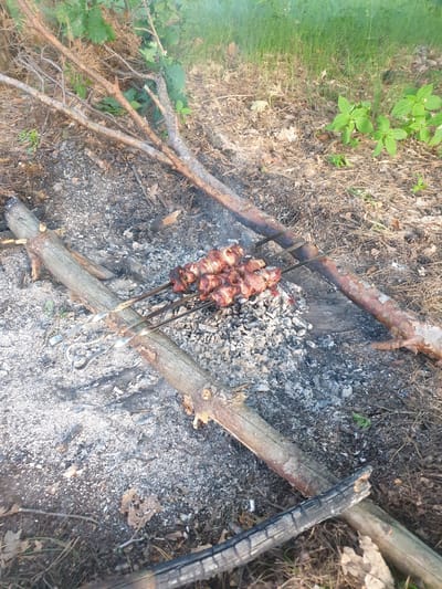 The image depicts an outdoor scene in Obukhiv, Ukraine, likely a wooded area or park, where meat is being grilled over a campfire. The foreground shows remnants of a fire, including ashes and charred wood. Several pieces of wood are arranged to form a makeshift grill. Metal skewers threaded with pieces of meat are suspended over the hot embers. The meat appears to be cooked, with some pieces showing browning and charring. Smoke gently rises from the grill, indicating that the cooking process is ongoing. The surrounding environment consists of dry leaves, pine needles, and sparse green vegetation. The lighting suggests it is daytime, possibly late afternoon or early evening. There are no people visible in the image.