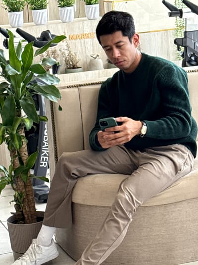 A man in a green sweater and khaki pants reclines on a beige sofa, gazing down at a green phone in his hand. He wears a gold watch. A potted plant with long, green leaves sits near the sofa's base. In the background, there are several small white potted plants on a wall, with the word "MARKET" visible. Part of an exercise bike is also present in the image.