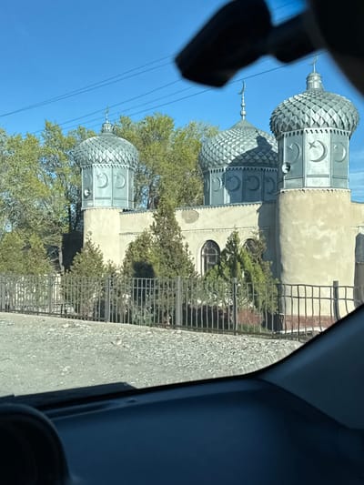 The image, taken from inside a vehicle, captures a structure with three silver domes, each adorned with a crescent moon and star symbol, indicative of Islamic architecture. The domes are connected to a beige building, partially obscured by trees and a black metal fence. The sky is clear and blue. The location is inferred to be in Chyrpykty, Kyrgyzstan.