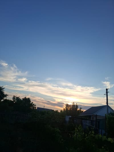 The image depicts an outdoor scene, likely in Kropyvnytskyi, Ukraine, during what appears to be sunset or sunrise. The sky is a prominent feature, transitioning from a clear, deep blue on the left to a sky filled with wispy clouds colored with hues of orange, pink, and yellow on the right, indicating the sun is low on the horizon.
In the mid-ground and extending into the right side of the frame, there are elements of a rural or semi-rural setting. A dark silhouette of trees and foliage frames the scene, suggesting a natural landscape. A portion of a building, possibly a house with a light-colored roof and a darker facade, is visible amongst the trees. Electrical wires stretch across the sky, supported by a utility pole, adding a man-made element to the natural scenery. The lower portion of the image is dominated by dark shadows, indicating the light source is low and casting long shadows.
There are no visible people or specific activities taking place. The overall mood is tranquil and serene, enhanced by the soft light of the setting or rising sun and the quiet, natural surroundings. The image captures a moment of natural beauty and the peacefulness of a rural evening or morning.