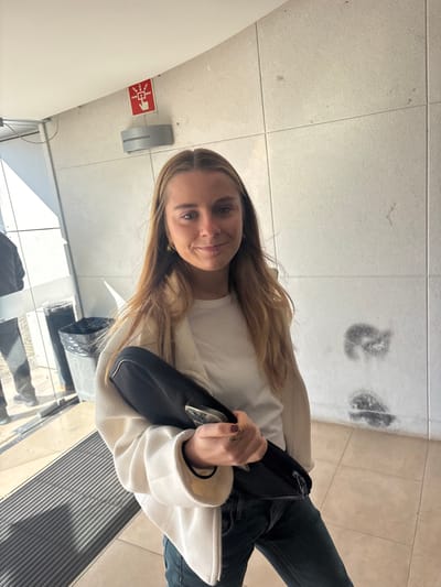 This image captures an indoor scene, located in the city of Porto Salvo, Portugal, featuring a young adult female subject and various environmental elements.

**People:**
The primary subject is a female with long, light brown hair, appearing to be in her twenties, positioned centrally and looking directly at the camera with a slight smile. She wears a white crew-neck t-shirt, an open cream or light beige fleece jacket, and dark blue jeans. Small gold-colored hoop earrings are visible. Her left hand is holding a black rectangular zippered case or bag, and a smartphone with a clear case is resting atop it. A ring is visible on her left ring finger. In the background, to the far left, a partial reflection of another individual in dark clothing and shoes is visible in a glass surface.

**Objects and Environment:**
The scene is set in what appears to be a well-lit hallway or entrance area of a building. The walls are composed of light-colored, possibly beige or off-white, square tiles with visible grout lines. On the wall above the primary subject's head, to the left, a red emergency pull station sign with white pictograms (an arrow pointing to a hand and vertical lines, with text "1982-1983") is mounted. Below this sign, a grey, curved wall-mounted light fixture is present. The right wall displays several dark, irregular smudges or marks, particularly two prominent ones on the lower section. The floor is covered with light-colored, possibly beige, square tiles. To the left, a glass door or partition is visible, reflecting sunlight and the partial figure of another person. A dark, cylindrical trash bin with a liner is visible through this glass. A rectangular metal grate is integrated into the floor near the base of the glass partition.