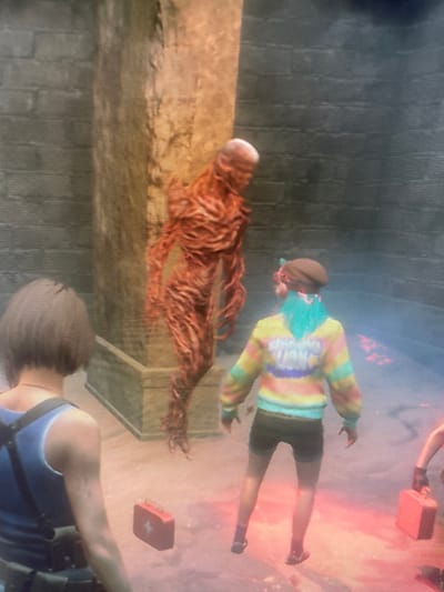 In a dimly lit, possibly subterranean environment, an elongated, skinless creature with a bald head lies on a ledge. A character in a colorful sweater stands beside the creature. Another character with short hair, wearing a blue top, is in the foreground. A red first-aid kit floats above the second character. The perspective of the image is from a third-person point of view. The image appears to be a screenshot from a video game.