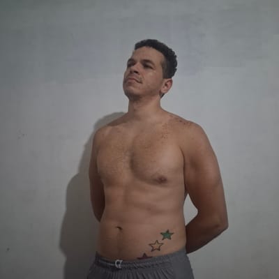 An adult male stands shirtless against a light-grey wall in what appears to be an indoor setting in Wilde, Argentina. He has short, dark, curly hair, stubble, and numerous small, dark brown macules distributed across his chest, shoulders, and neck. His arms are positioned behind his back, obscured from view. He is wearing grey athletic shorts with a white drawstring. On his left lower abdomen/hip, there is a tattoo depicting three outlined stars in an ascending diagonal arrangement: a reddish-brown star, a yellowish-orange star, and a green star. The man is looking upwards and slightly to his right. A soft shadow of his left side is cast onto the wall behind him.