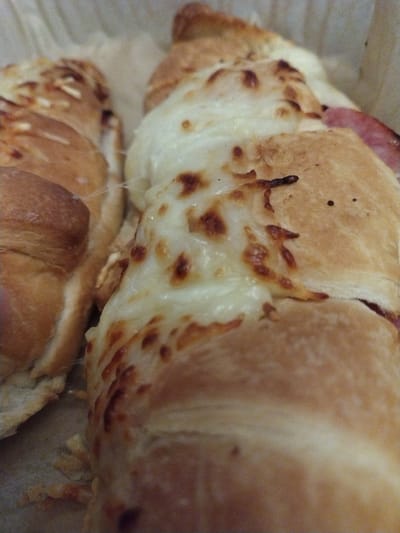 The image displays a close-up, slightly angled view of multiple savory baked pastries, resembling ham and cheese croissants or similar rolls, contained within a light-colored, possibly paper or parchment-lined, tray or container. This scene is located in Crauthem, Luxembourg. The pastries possess a golden-brown, flaky crust. Each is covered with a substantial layer of melted white cheese, which exhibits areas of golden-brown caramelization and bubbling, indicating it has been recently baked or heated. Portions of pinkish cured meat, consistent with ham, are visible beneath the cheese and within the pastry structure, particularly on the item positioned to the right. The items are arranged in parallel, partially overlapping, and fill the majority of the frame. No individuals are present, and no actions or interactions are depicted. The lighting is diffused, suggesting an indoor environment.
