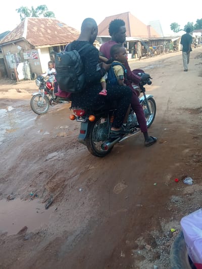 This image shows a family riding a motorcycle down a muddy, unpaved road in Kagini, Nigeria. A man wearing a maroon shirt and dark, patterned pants is driving the motorcycle, while a woman in a black, polka-dotted dress sits behind him, holding a young child. The child, wearing a yellow shirt and pink sandals, is seated in front of the woman. A second, younger child, with pink sandals, is visible behind the driver. Both children appear to be seated on the motorcycle's passenger seat or directly on the laps of the adults.
The scene appears to be set in a rural or semi-urban area, with buildings and market stalls lining the road. The buildings have corrugated metal roofs, and some appear to be shops. The road is in poor condition, with puddles of water and mud indicating recent rain or poor drainage. In the background, another motorcycle is parked, and a person is walking away from the camera.
The time of day appears to be late afternoon or early evening, suggested by the warm, diffused light and long shadows. The weather seems to be overcast or humid. The overall atmosphere is one of everyday life in a community where motorcycles are a primary mode of transportation. No explicit text is visible in the image, though posters or signs might be present on the buildings in the background, too small to read.
