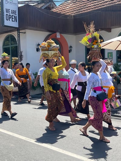 A street in Ubud, Indonesia features a procession of Balinese women. They are dressed in traditional Balinese clothing and carrying offerings on their heads. The procession is passing by a restaurant called Basilico Fusion Cuisine, which has a sign indicating that it is open. The women are walking in a line, moving from the bottom to the top of the frame, and their faces exhibit focused attention. The photograph is oriented vertically, and the subjects are positioned parallel with the horizon.