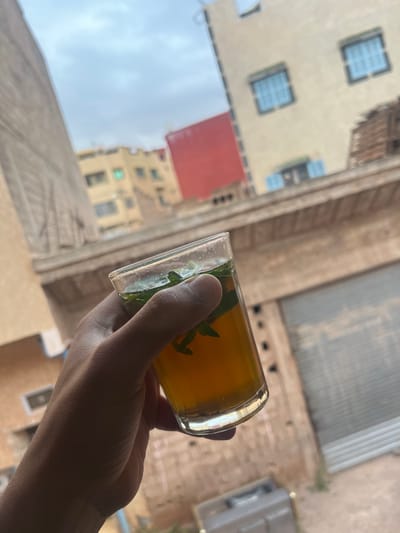 A hand holds a glass of tea with mint sprigs, with the city of Ouaouizert, Morocco, visible in the background. The hand is light-skinned and slightly shadowed. The glass is cylindrical and appears to be made of clear glass. The tea is a light amber color, and green mint sprigs are visible inside the glass. Outside the window, buildings with windows, a red roof, and a gray corrugated metal roof can be seen. The sky is overcast. The window frame is made of weathered wood. The perspective is looking slightly upward toward the buildings.