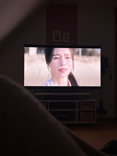 The image shows a person's legs in the foreground, obscuring the lower part of the scene. A large television screen is in the mid-ground, displaying a close-up of a woman's face. She has dark hair, is wearing a light-colored garment, and has a subtle smile. Behind the television, a white shelf unit with two woven baskets is visible. To the right of the television, a dark-colored vacuum cleaner stands vertically. The background appears to be an indoor setting with dimly lit walls. The scene is not located in Breisach am Rhein, Germany; no elements in the image indicate this specific location.