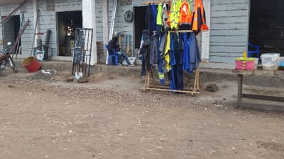 A street scene presents a row of buildings with corrugated metal siding. Work clothes hang on a wooden rack. A person sits in a blue plastic chair, looking at a mobile phone. Adjacent is a table with colorful containers. Debris litters the ground, and a motorcycle stands to the left of the image.