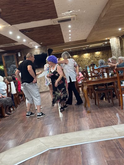 The image depicts a group of people inside what appears to be a restaurant or a banquet hall. The setting features a wooden floor and ceiling with decorative elements. In the foreground, a man in a black t-shirt and grey shorts is interacting with a woman wearing a vibrant purple wig and a floral dress. Another woman in a white top and black pants is also visible. Several other individuals are seated at tables in the background, some of whom are eating or conversing. The ambiance suggests a social gathering or event. The location is identified as Sredets, Bulgaria.