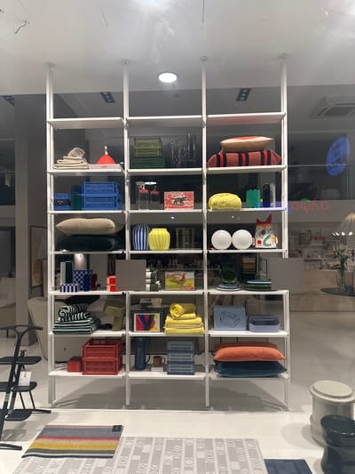 The image captures a vibrant display inside what appears to be a modern retail store or showroom, featuring a large, white, grid-like shelving unit filled with various home goods and decorative items. The perspective suggests the photo was taken through a glass window or partition, as reflections of the interior and text are visible.

The central shelving unit is organized into numerous square and rectangular compartments, each holding carefully arranged products. These include a variety of pillows in solid colors (beige, dark green, orange, light blue) and striped patterns (red and black, light yellow and white, green and white). Decorative vases, some striped (yellow and white, blue and white) and others solid, are also displayed. Storage crates in green, blue, and red are interspersed throughout the shelves, alongside stacks of folded towels (yellow, cream, striped), bowls, small sculptures (including a colorful fish-shaped one), books or magazines, and framed abstract art.

On the floor to the right, two rugs are visible: one light gray with a repeating geometric pattern, and another with prominent horizontal stripes in multiple colors including red, yellow, blue, and green. In the reflections on the glass, parts of the store's interior are visible, including what seems to be a table with chairs, other products, and ceiling lighting fixtures. Visible reversed text in reflection includes "ayda" and a phrase possibly reading "for your future." The lighting is bright and artificial, illuminating the products clearly. The overall aesthetic is clean, contemporary, and colorful, showcasing a curated collection of household and decor items.