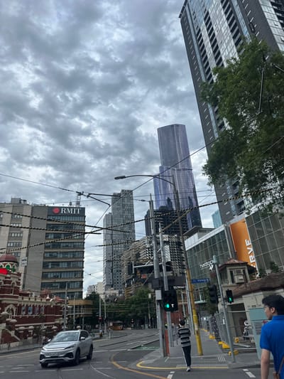 This image captures a wide-angle urban street scene at the intersection of Victoria Street and Swanston Street in Melbourne, Australia, under a heavily overcast sky.

In the left mid-ground, a multi-story building featuring the red "RMIT" logo on its rooftop dominates, displaying a facade of horizontal banding and numerous windows. To its right, a series of modern skyscrapers rise, including the prominent, slender, purple-tinted Aurora Melbourne Central tower, and another multi-patterned grey residential high-rise. Overhead, a dense network of black tram and power lines, supported by multiple utility poles, crisscrosses the frame, partially obscuring the buildings and sky.

On the street level, a silver SUV (license plate "1G9 4ES") is captured in motion, initiating a right turn from a traffic lane that includes tram tracks. Further down the street, a red tram is faintly visible. Multiple traffic light poles are present; one in the right mid-ground displays a green 'T' signal for trams and a green pedestrian signal.

Two individuals are visible: one person in a blue shirt is partially seen from behind in the far right foreground, walking away from the camera. Another person, wearing a black and white horizontal striped shirt with dark pants and white shoes, walks on the sidewalk in the mid-ground, moving away from the viewer.

On the right side of the frame, parts of commercial buildings are visible, including one with a large orange "ERVE" sign and another with "hardrock" signage, alongside an older, smaller, light-colored building with a distinctive terracotta-colored roof. Green tree foliage is present on the extreme right, partially obscuring a building. The overall environment is a busy city intersection with active pedestrian and vehicular traffic.