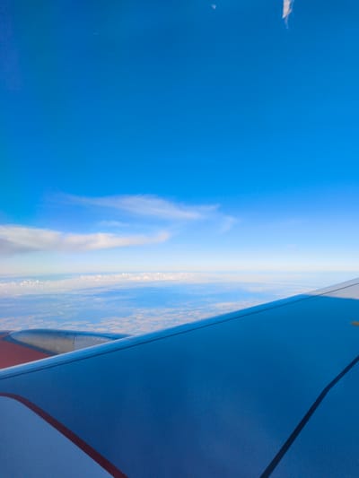 The image is an aerial view from an airplane window, looking down at the Earth and up at the sky. The lower portion of the frame is dominated by the wing of the airplane, which is painted a deep blue with a red stripe along its edge. To the left, a portion of the airplane's engine nacelle is visible, appearing metallic and cylindrical.

Above the wing, a vast expanse of land stretches to the horizon. The land appears to be agricultural, with fields of varying shades of brown and yellow, suggesting a cultivated landscape. A thin line of clouds is visible on the horizon, separating the land from the sky.

The sky above is a vibrant, clear blue, with a few wispy white clouds scattered across it. The overall impression is one of flight and vastness, with the airplane providing a human element within the natural landscape.

There is no information in the image that indicates it is specifically located in Schipkau, Germany.
