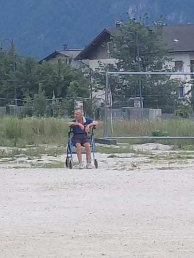 An elderly man sits on a blue walker, facing right, with his arms resting on the handles. He is wearing a blue polo shirt and dark shorts. He is outdoors, on a light-colored, gravelly surface. Behind him, there is green grass, trees, and buildings. The background shows a large, green hill or mountain. The scene is likely in Levico Terme, Italy, given the presence of buildings and a natural mountainous backdrop.