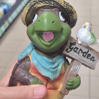 A close-up, slightly upward angled shot features a decorative garden statue of a smiling cartoon turtle wearing a straw hat and a light blue scarf. The turtle is holding a small wooden sign that reads "Garden" in black cursive. A small white and green bird is perched on top of the sign, facing left. The statue is held in a person's hand, with the thumb and index finger visible. The background is blurred and appears to be an indoor setting, possibly a store, with rows of products on shelves in the upper left corner. The overall impression is a whimsical and friendly garden decoration. The image was taken in Kokshetau, Kazakhstan.