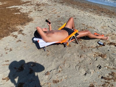 The image shows a person lying on a beach chair on a sandy beach. The person is shirtless and wearing black swim trunks. They are holding a smartphone and appear to be looking at it. A white towel is placed beneath the chair. Orange flip-flops are visible on the sand to the right of the chair, as well as a dark can. The beach is covered in sand with a significant amount of dried seaweed scattered across it. In the background, the ocean waves are visible. The sun appears to be high in the sky, suggesting it is midday, and the weather is sunny. The shadow of the person taking the photo is prominently cast on the sand in the foreground. The location is Estepona, Spain.