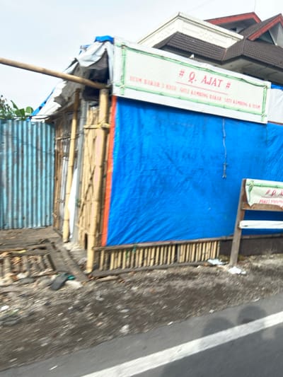 The image shows a building under construction, located in Kecamatan Margahayu, Indonesia at latitude -6.976, longitude 107.564. The front of the building is covered with a blue tarp, secured with a wooden frame. The building is made of concrete and has a sign that reads "Q. AJAT" in red text. The sign also mentions "D. BAHAN 2 RINC" in smaller text. The building is located on a dirt road with a white painted lane. The image is captured from a moving vehicle, resulting in blurry details. The surrounding environment is also blurred, making it difficult to discern any other objects or structures. The image shows a construction site in a residential area of Kecamatan Margahayu.