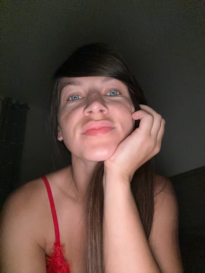 A woman with light skin and blue eyes rests her chin on her hand. She has long, dark brown hair that falls over her shoulder. She is wearing a red top with lace trim. The background is dark and out of focus, with a hint of a wall on the left side of the image. The lighting is dim, suggesting an indoor setting, possibly a bedroom or living area. The photograph appears to be a selfie, taken from a close-up perspective, focusing on the woman's face and upper body. The specific location in Donji Pobori, Montenegro, is not visually discernible from the provided image and its crops.