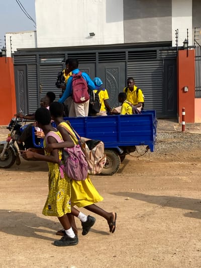 A group of people are traveling by motorcycle with an attached blue trailer. People are riding in the trailer wearing yellow shirts or dresses. The motorcycle is on a dirt road in front of a building with a gray gate. Some of the people are wearing backpacks.