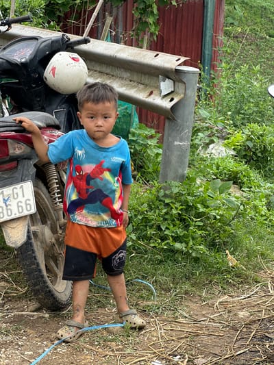 A young East Asian male, appearing between four and seven years old, stands outdoors in Mù Cang Chải, Vietnam. He has dark, short hair and dirt smudges on his forehead, cheeks, and lower legs, where faint temporary tattoos are also visible. He is dressed in a blue short-sleeved t-shirt featuring a red and blue Spiderman graphic with "MARVEL" and "SPIDER-MAN" text, orange and black shorts with a white stylized graphic on the right outer thigh, and soiled grey-green Crocs-style clogs. A light blue ribbon or string is present on the ground around his left foot.

The boy's left hand rests on the rear seat of a dark-colored Honda scooter-style motorcycle, which displays a white license plate with the registration "G1 86.66" on its muddy rear fender. A white helmet with a red Honda wing logo and handwritten text, including "XINH", hangs from the motorcycle's handlebars. Behind the boy and motorcycle, a weathered, corrugated metal guardrail with a white reflective strip is affixed to a grey cylindrical post. Further back, a red-brown corrugated metal wall with a vertical green strip is partially visible behind dense green foliage. The ground is uneven, composed of dirt and mud, with scattered dry leaves, twigs, and various green plants and grasses. A yellow-brown plastic object is partially obscured by grass on the right side of the frame.