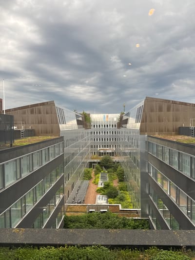The image shows a view of the UN City (United Nations City) in Copenhagen, Denmark, from a higher vantage point. The foreground features a concrete barrier and dense green foliage. The main subject of the image is the distinctive architecture of the UN City complex, characterized by its modern, angular buildings with extensive glass facades and green roofs. The buildings are arranged around a central courtyard filled with trees and greenery, with walkways and bicycle racks visible. The sky is overcast with dark, dramatic clouds, creating a moody atmosphere. Reflections of the sky and surrounding structures are visible in the glass surfaces of the buildings. A communication tower can be seen in the distance. There are no people visible in the image.